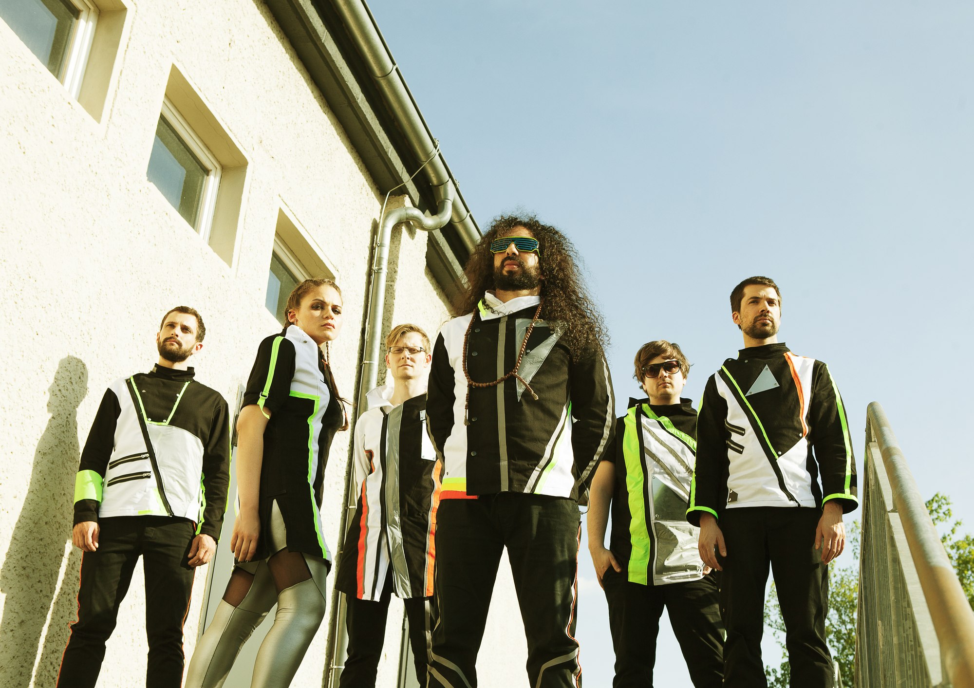 The six members of Out Of Nations standing together outdoors in matching futuristic stage costumes, under a clear blue sky