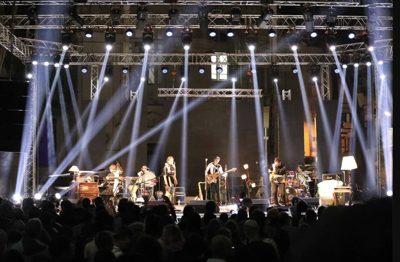 Out Of Nations performing on a large outdoor stage under white light beams, in front of a big crowd